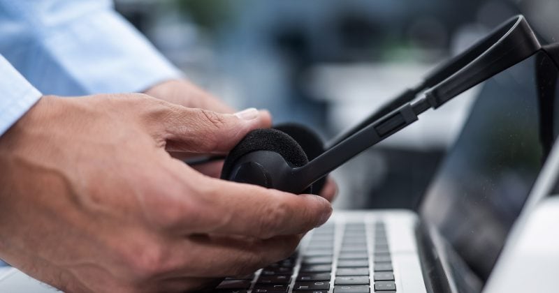 A person places a black customer service headset on a laptop keyboard, with one hand holding the headset and the other on the laptop. The background is blurred.