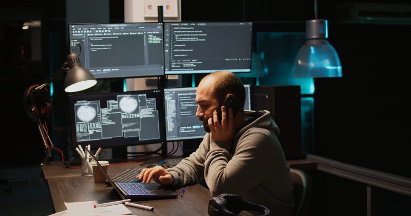 A person sits at a desk with a laptop and multiple monitors displaying code, discussing website security on the phone in a dimly lit office.