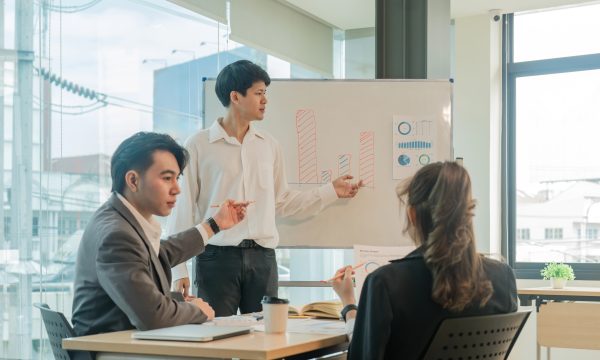 Three people in a meeting room; one person stands and presents bar charts on a whiteboard while two others sit at a table and listen.