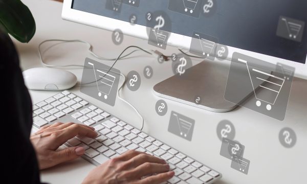 Hands typing on a keyboard in front of a monitor, with shopping cart and currency icons digitally superimposed, representing online shopping.
