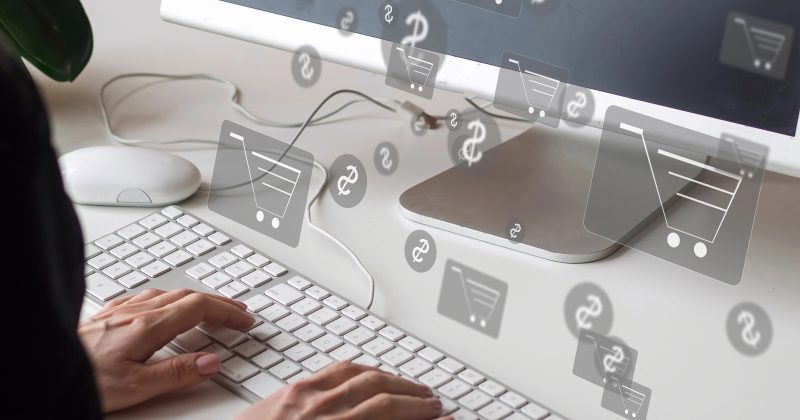 Hands typing on a keyboard in front of a monitor, with shopping cart and currency icons digitally superimposed, representing online shopping.