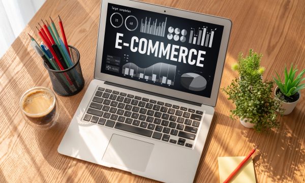 A laptop on a wooden desk displays graphs and the word E-COMMERCE, hinting at WordPress tools. Nearby are a cup of coffee, colored pencils, a plant, a notepad, and a red pencil.