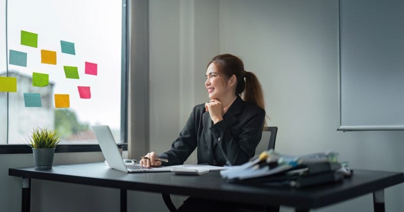 Woman sitting at a desk with a laptop, looking out the window; colorful sticky notes on the glass and papers spread on the desk, perhaps brainstorming ways to attract talent using the best WordPress plugins.