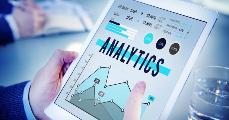A person uses a tablet displaying analytics data, including graphs and charts from WordPress Google Analytics Plugins, while seated at a table with papers and a glass of water nearby to track site performance.