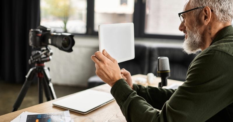 Older man with gray hair and beard holds a tablet while sitting at a desk, recording a video in front of a camera and microphone, sharing tips on how to maximize engagement using a gated content plugin for WordPress in a modern office setting.