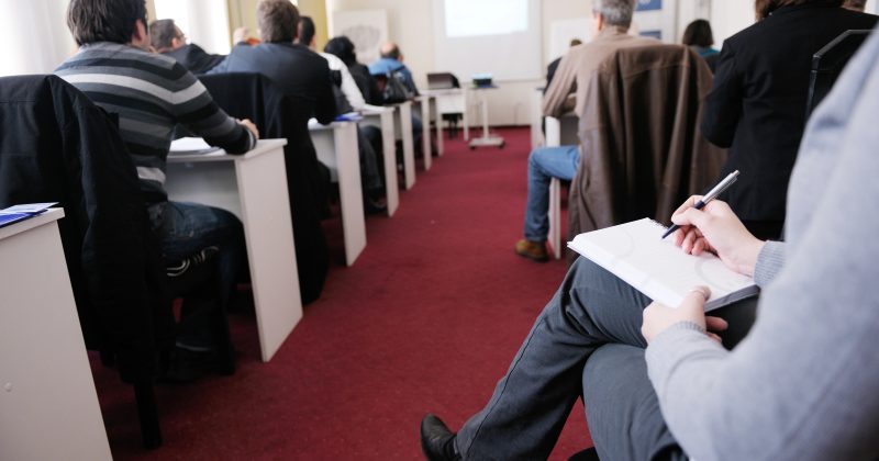 People seated in a classroom, taking notes and facing a projector screen during a presentation or lecture.