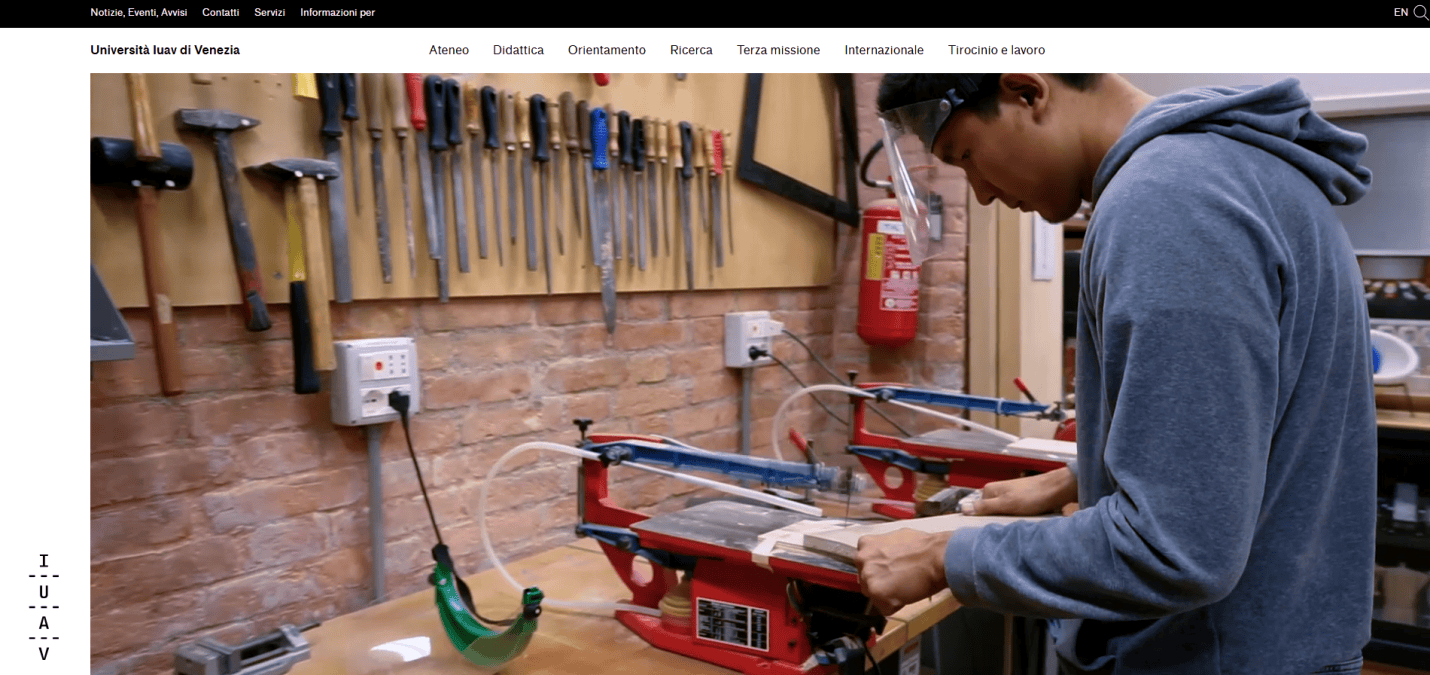A person wearing safety goggles works with a scroll saw in a workshop. Various hand tools hang on the wall, and a fire extinguisher is mounted nearby—a scene reflecting the practical inspiration behind 2025 education website design.