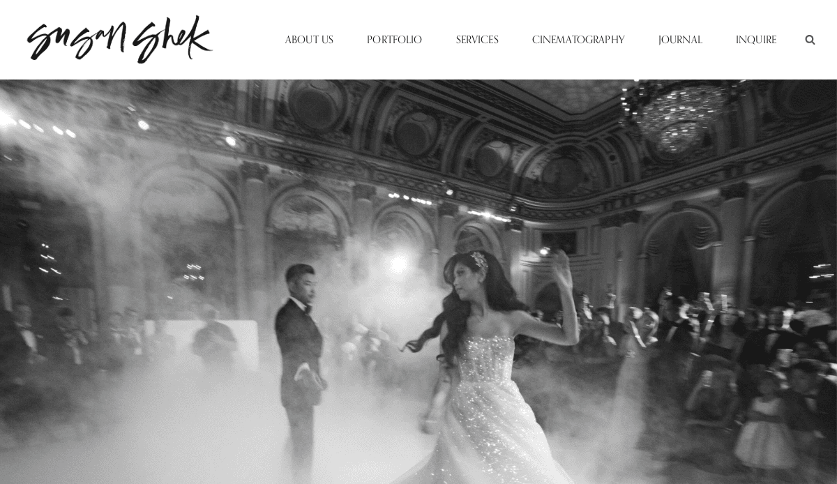 A black-and-white photo of a bride and groom dancing in a grand ballroom with high ornate ceilings, chandeliers, and guests watching amid swirling fog offers timeless wedding inspiration.