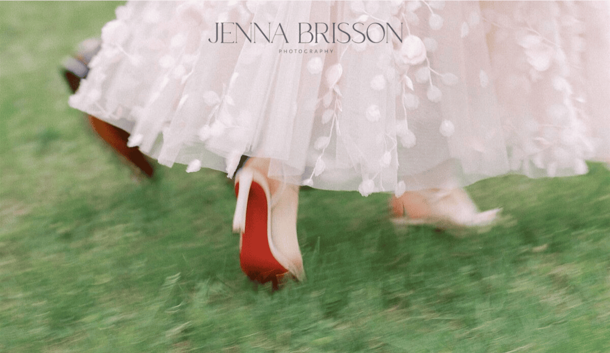 A close-up of two people walking on grass, featuring a flowing, delicate white dress with floral details and red-soled high heels. The image is soft-focused, perfect for wedding website designers, with the photographer’s watermark at the top.