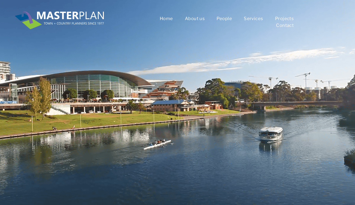 A modern riverside building with a curved roof sits beside a green lawn, showcasing elegant landscape design. A rowboat and a small ferry travel on the calm river under a clear blue sky. Landscaping website navigation text is visible at the top.