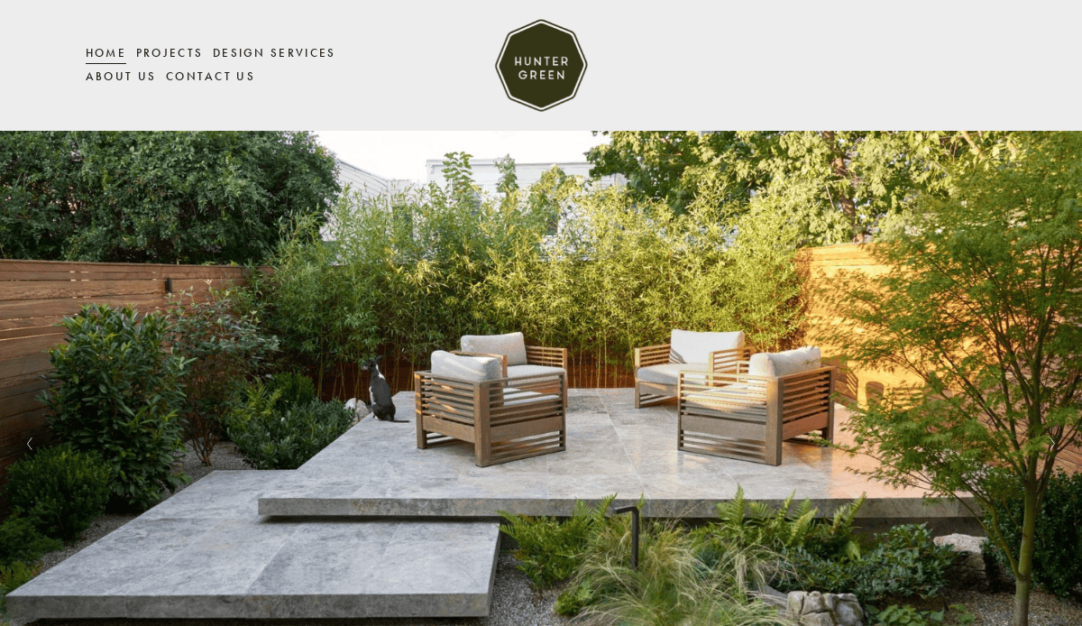 A modern garden patio with four wooden armchairs and white cushions sits on a stone platform, surrounded by lush greenery and a wooden fence—perfect inspiration for your next landscaping project. Hunter Green logo is at the top center.