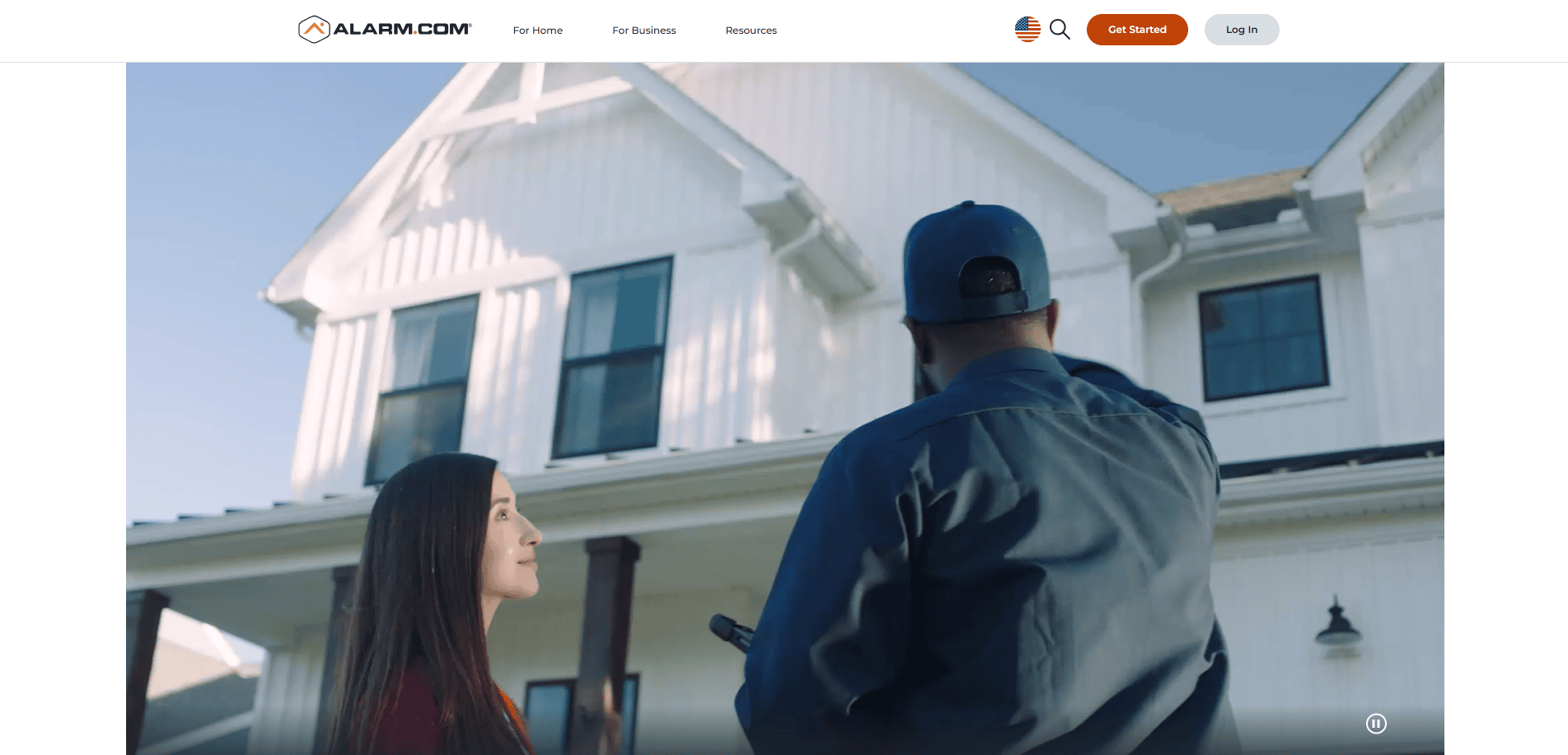 A woman and a technician stand outside a white house with black-trim windows. The technician points up at the house while the woman looks on. The Alarm.com logo and navigation menu are visible at the top of the image.