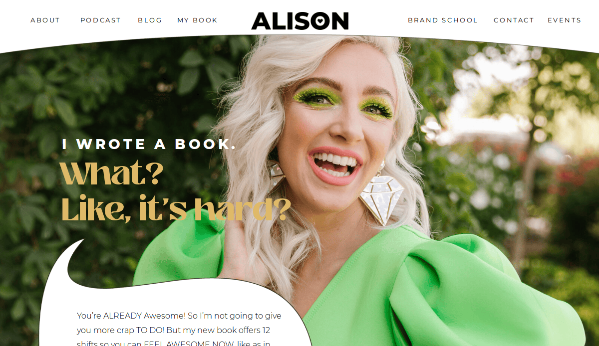 A smiling woman with platinum blonde hair and bright green eye makeup wears a vibrant green outfit and large geometric earrings, standing outdoors. Text says, “I WROTE A BOOK. What? Like, it’s hard?”.