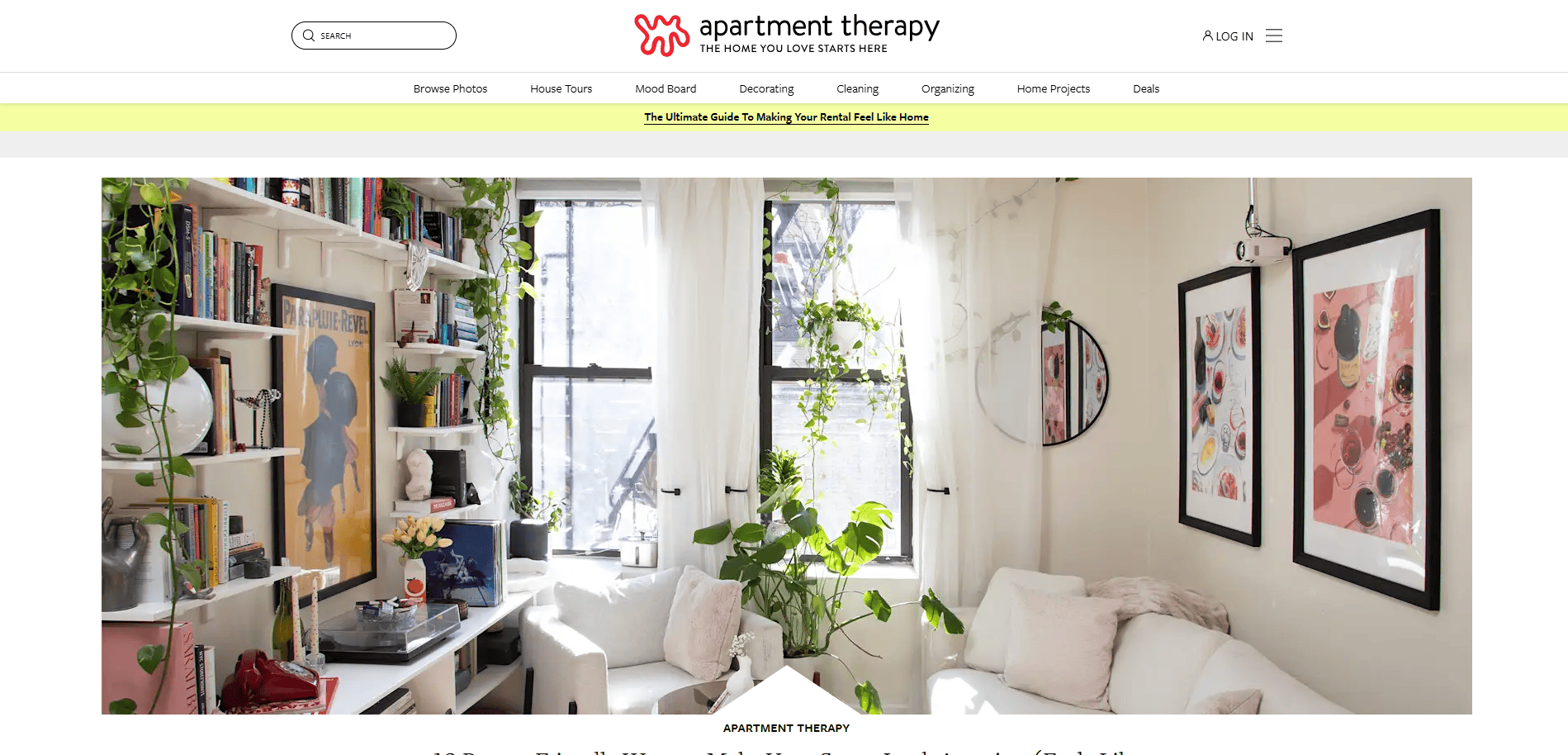A bright, cozy living room with white walls, a white sofa, plants on shelves and hanging, a window letting in sunlight, and colorful wall art. Books and decorations fill the shelves on the left.