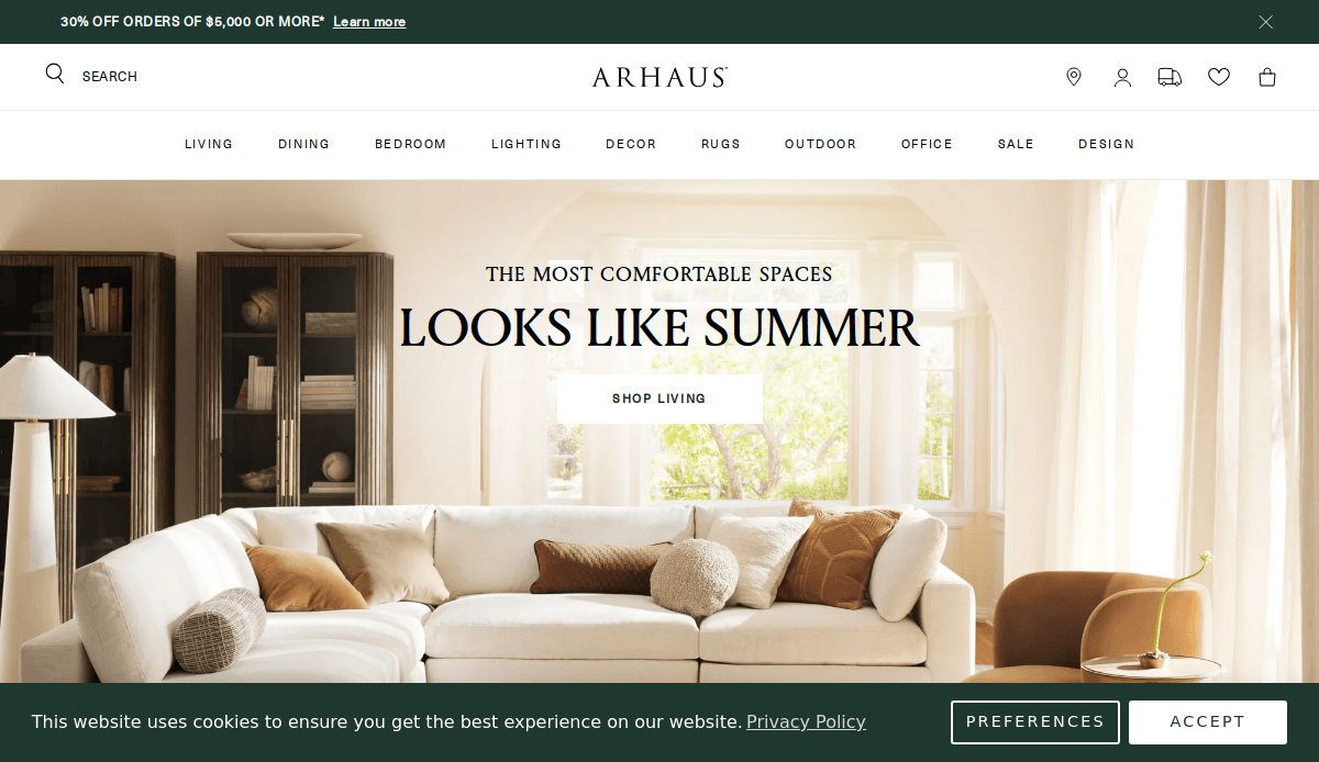 A stylish living room with a white sectional sofa, cozy pillows, and neutral décor under large arched windows. The text reads, The Most Comfortable Spaces Looks Like Summer with a Shop Living button.