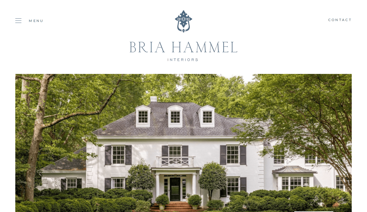 A stately white two-story house with black shutters, surrounded by lush green trees and manicured bushes, featured on the Bria Hammel Interiors website homepage.