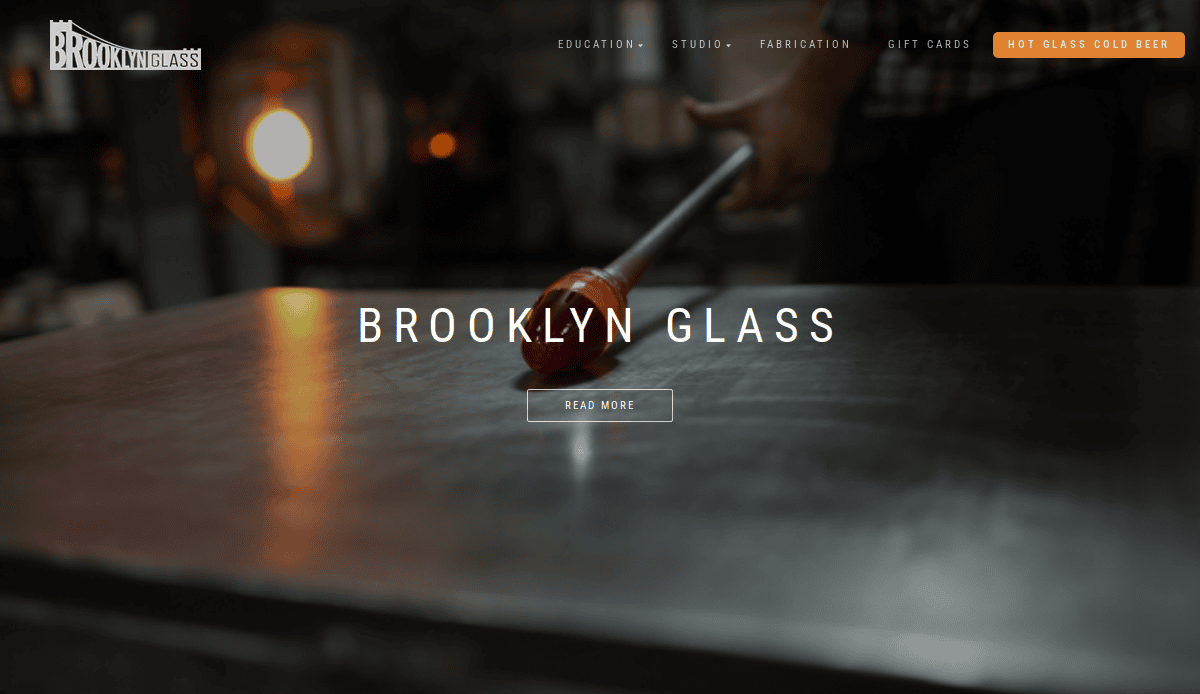 A person shapes hot glass on a metal table in a glassblowing studio, with the Brooklyn Glass logo, navigation menu, and “BROOKLYN GLASS” text overlayed on the image.