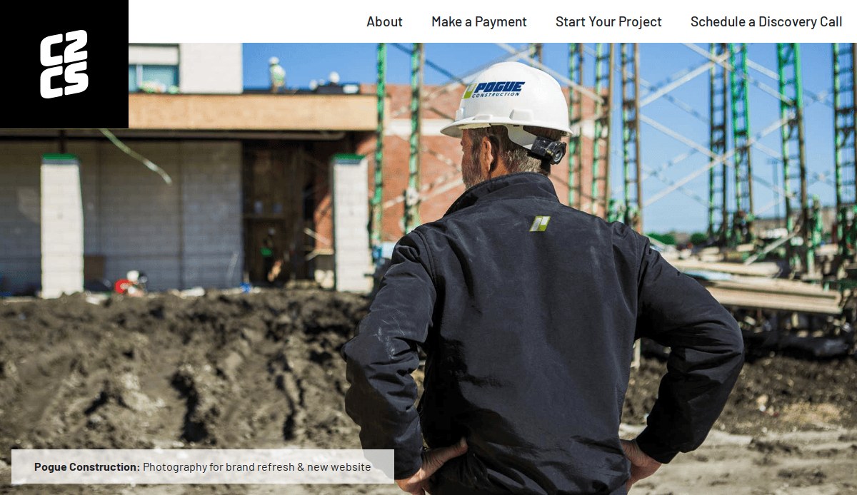 A construction worker wearing a hard hat and black jacket stands with hands on hips, facing a building under construction with scaffolding and dirt in the foreground.