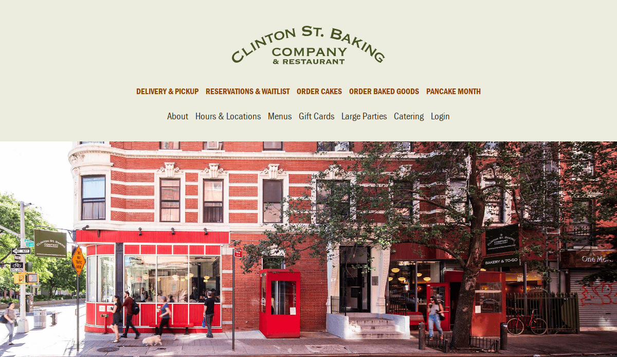Street view of Clinton St. Baking Company & Restaurant, a corner building with red and white exterior, large front windows, and outdoor seating surrounded by trees and neighboring businesses.