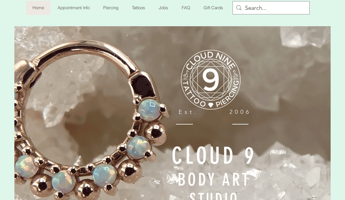 Close-up of a gold hoop earring with opal stones on a crystal background. The screen shows the Cloud 9 Body Art Studio website with navigation links and the studios circular logo.