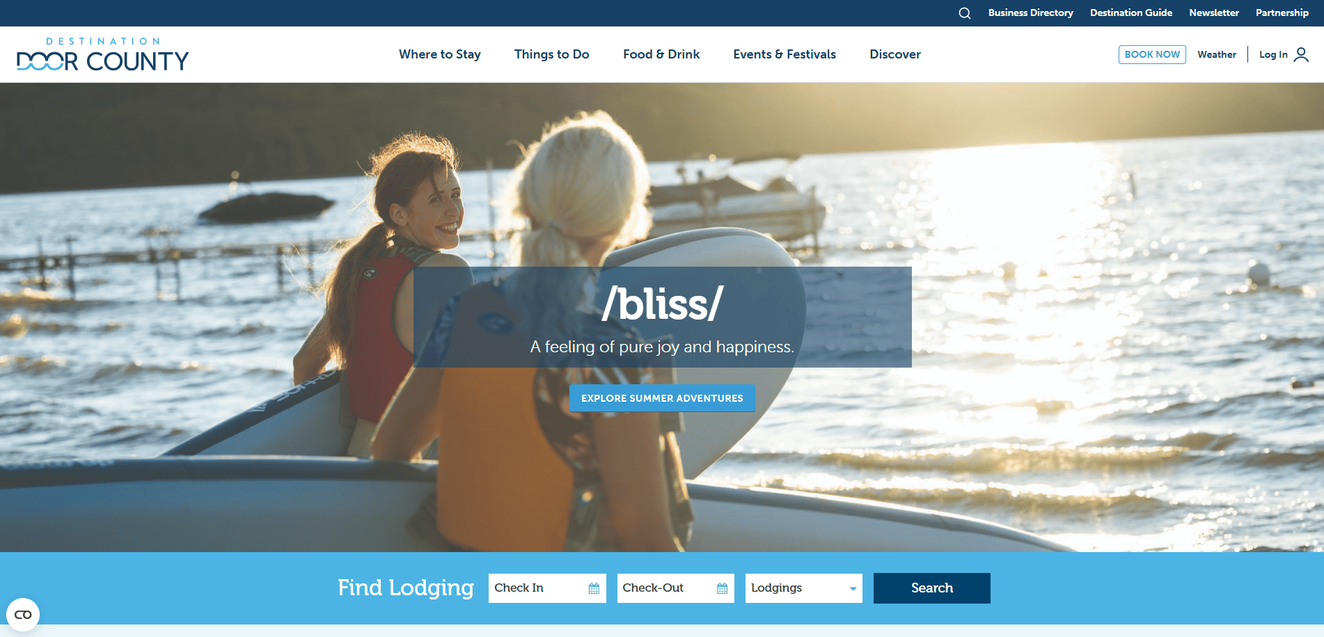 Two women in swimwear sit on stand-up paddleboards on a sunlit lake, smiling at each other. The water glows with sunlight and trees line the distant shore. Text reads, “/bliss/ A feeling of pure joy and happiness.”.