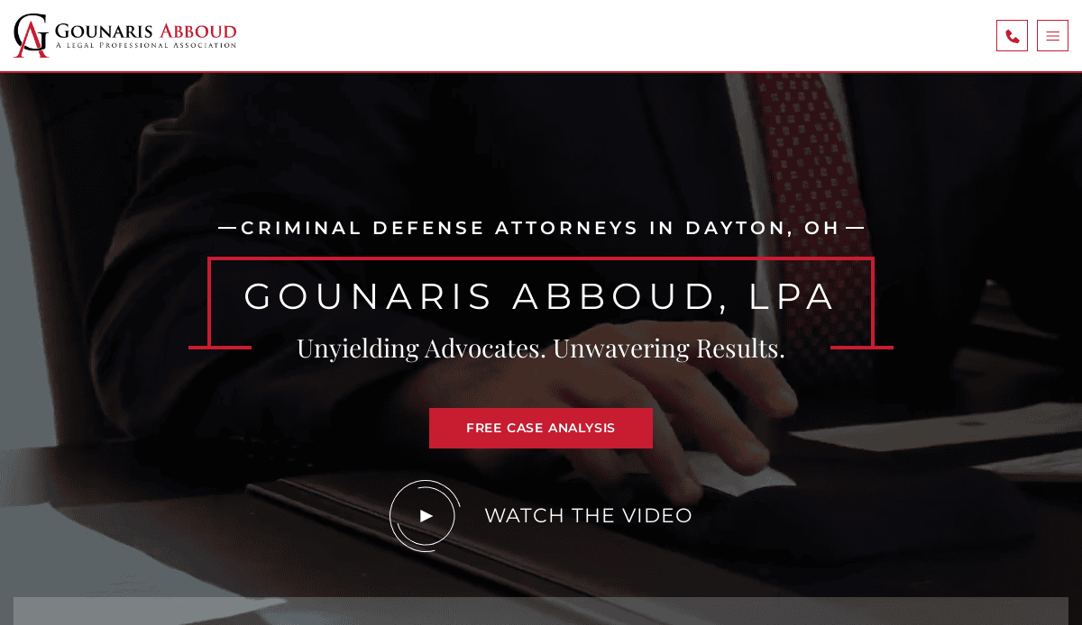 A person in a suit writes on a notepad at a desk. The website text reads: “Gounaris Abboud, LPA—Criminal Defense Attorneys in Dayton, OH. Unyielding Advocates. Unwavering Results. Free Case Analysis. Watch the video.”.