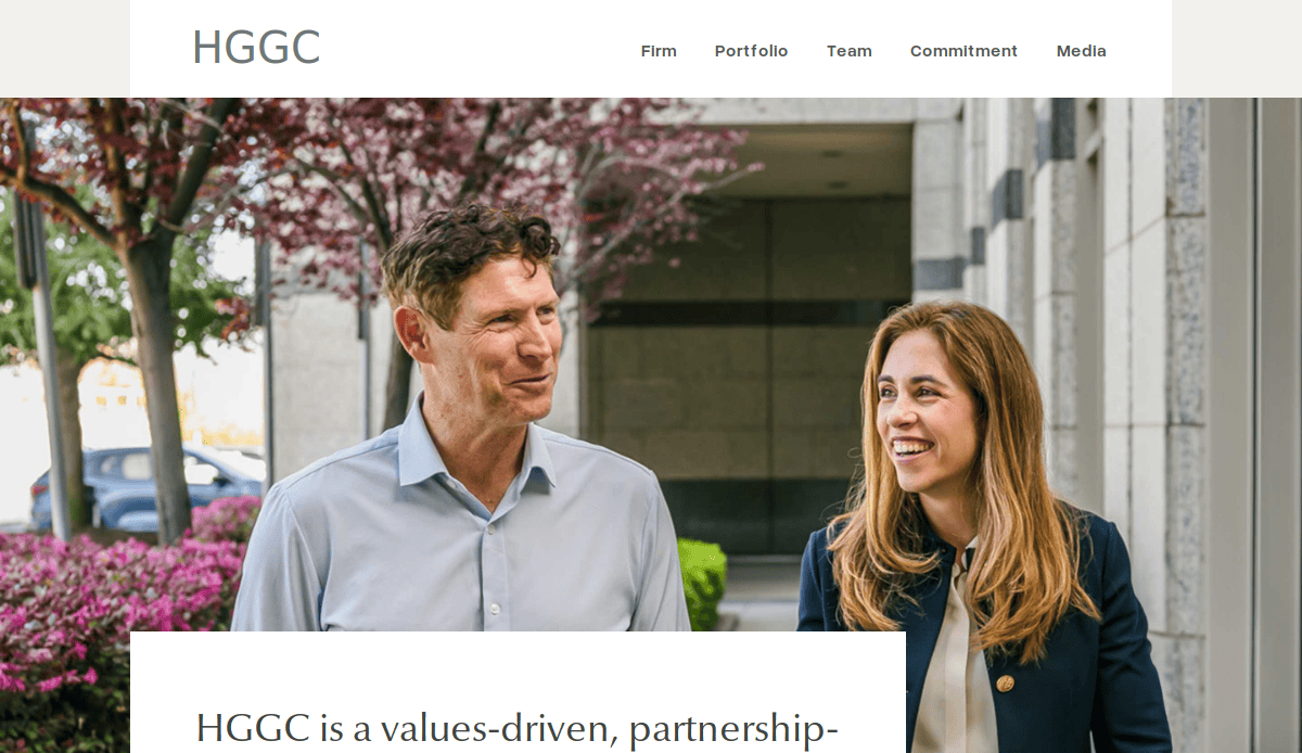 Two business professionals, a man and a woman, walk and talk outside an office building with pink-blossomed trees and greenery in the background. The HGGC logo and website navigation are visible at the top.