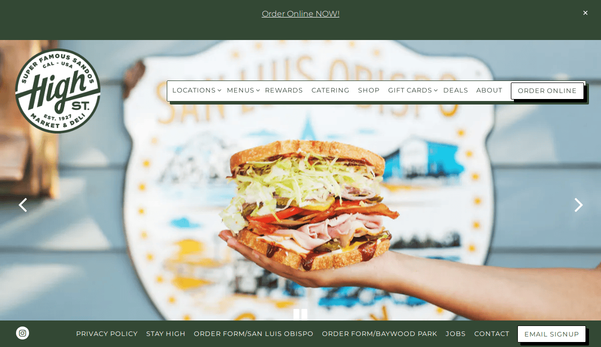 A hand holds up a large, stacked deli sandwich in front of a blurred High St. Market & Deli sign, which features the text “High St.” and “San Luis Obispo.” Menu and online ordering links are visible at the top of the image.