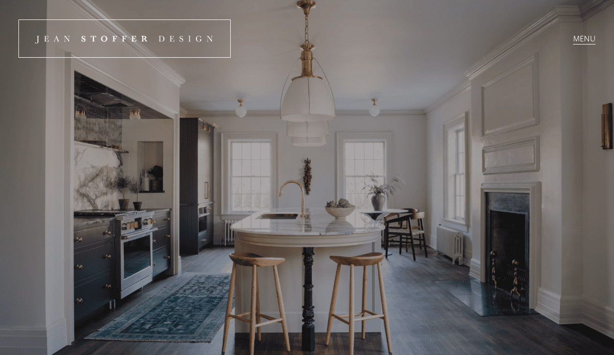 Bright, elegant kitchen with a central island, two wooden stools, brass fixtures, white cabinetry, dark stove, dining nook by windows, and a fireplace; “Jean Stoffer Design” text appears in the top left corner.