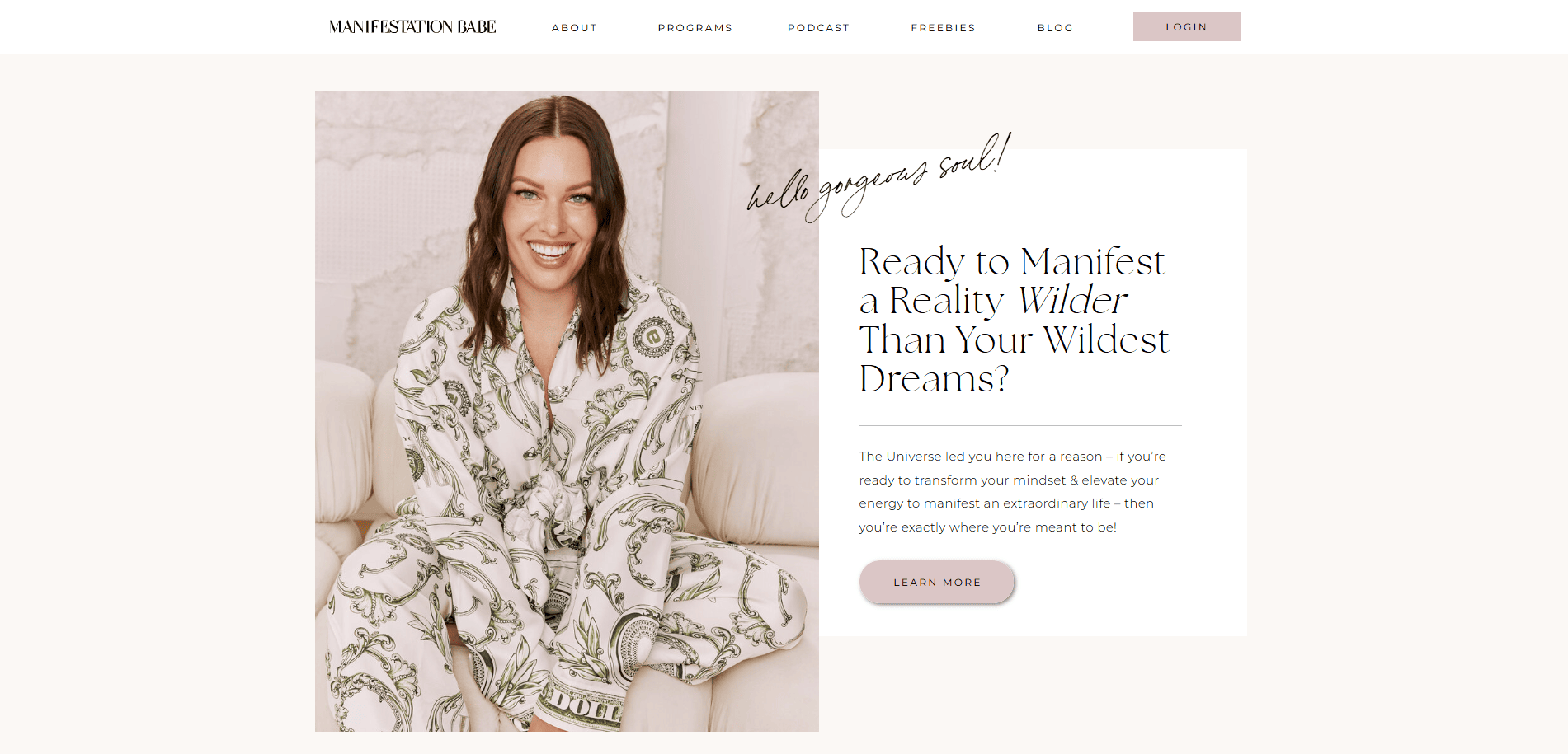 A smiling woman in patterned pajamas sits cross-legged on a couch. The text reads, “Ready to Manifest a Reality Wilder Than Your Wildest Dreams?” with a button labeled “Learn More.” The website is for Manifestation Babe.