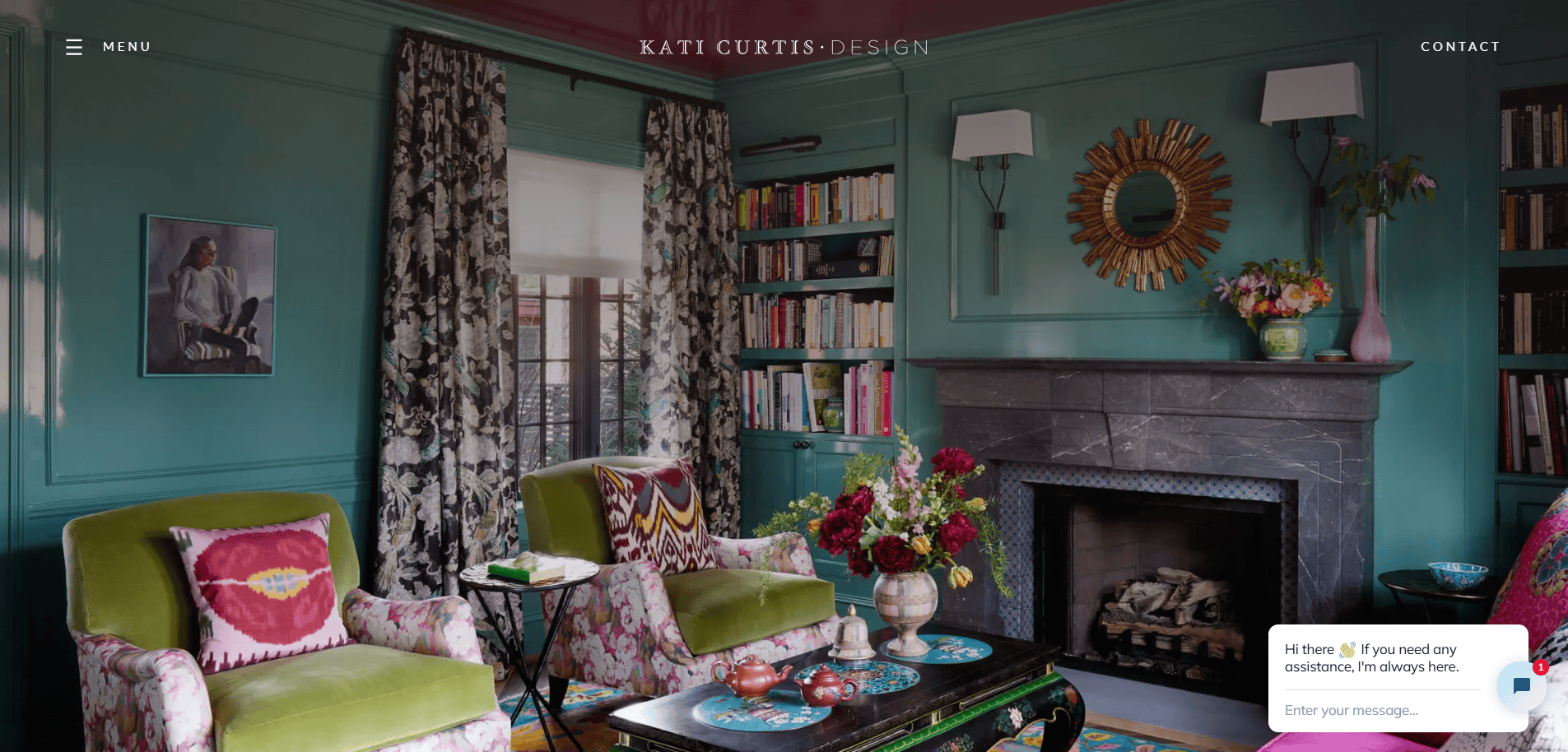 A stylish living room with green walls, floral curtains, velvet armchairs, a marble fireplace, bookshelves, a sunburst mirror, and a coffee table with flowers. A chat box is open in the bottom right corner.