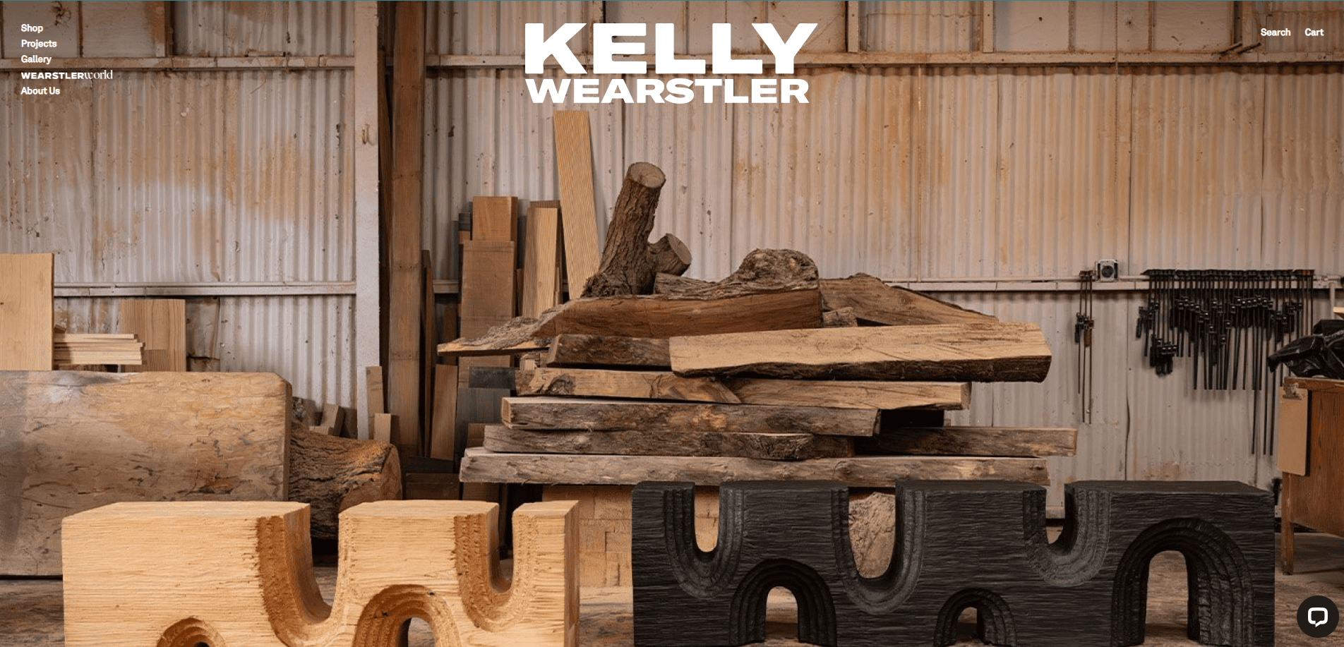 A workshop with stacked wooden slabs and sculpted wooden furniture pieces in the foreground. The text Kelly Wearstler is displayed at the top, overlaying the industrial-style workspace.