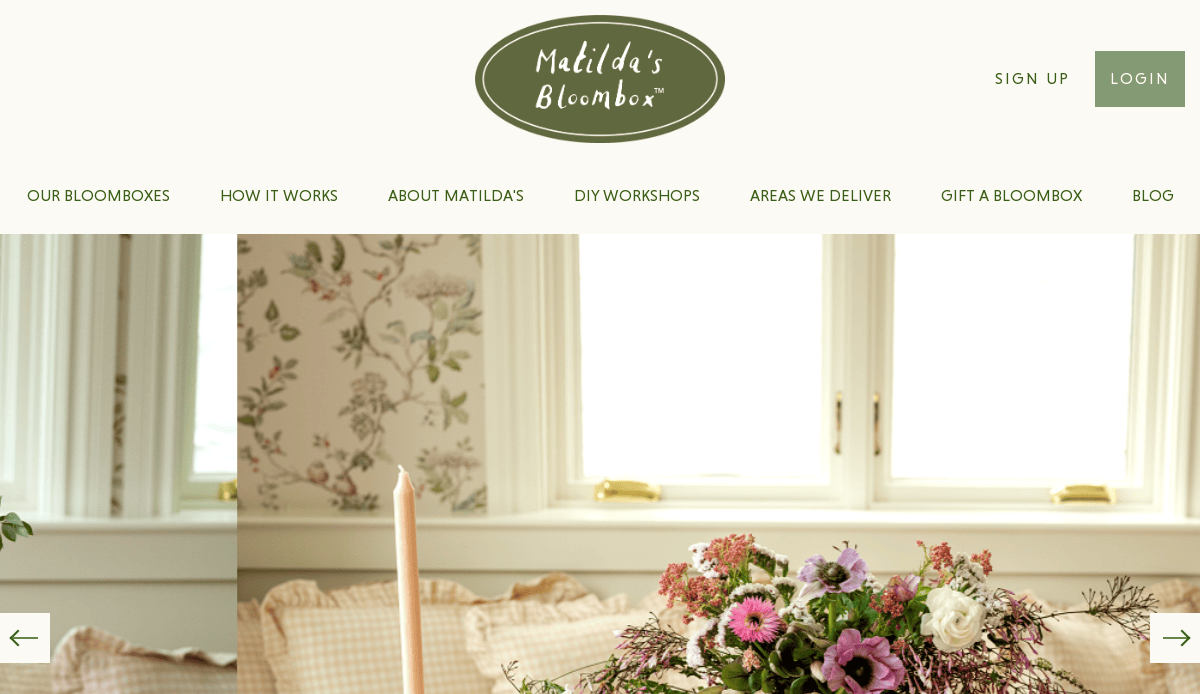 A website homepage for Matilda’s Bloombox featuring a floral arrangement on a table in front of two windows, with navigation links and options to sign up or log in at the top.