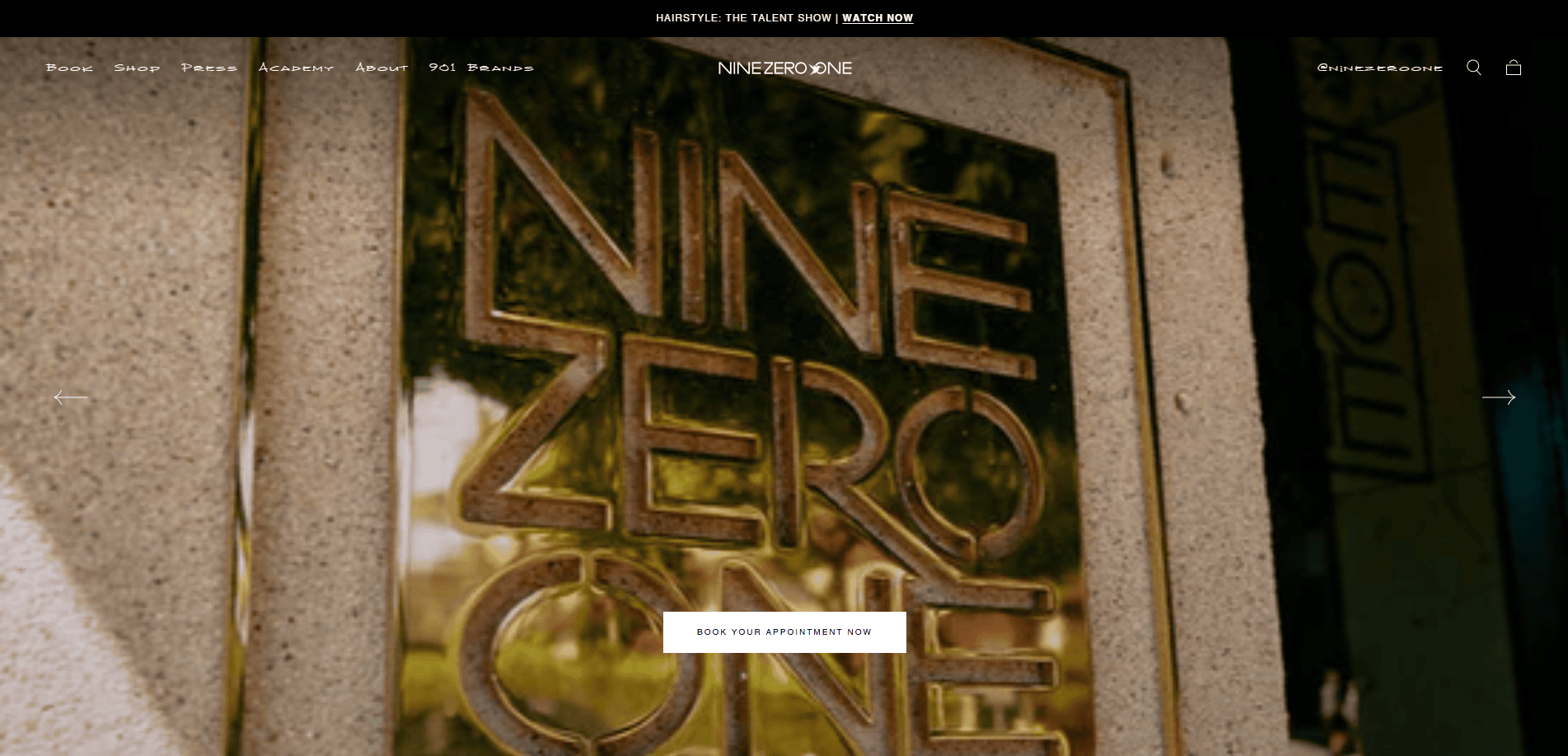 A close-up of a metallic sign reading NINE ZERO ONE mounted on a stone wall, with greenery and reflections visible in the background.