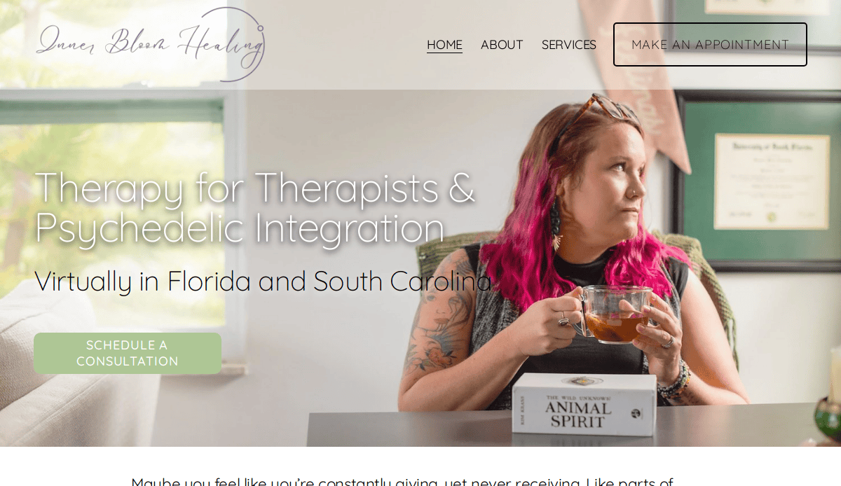 A woman with pink hair sits at a table holding a teacup, looking to the side. A deck of Animal Spirit cards is in front of her. Text on the image promotes therapy, psychedelic integration, and holistic web design services.