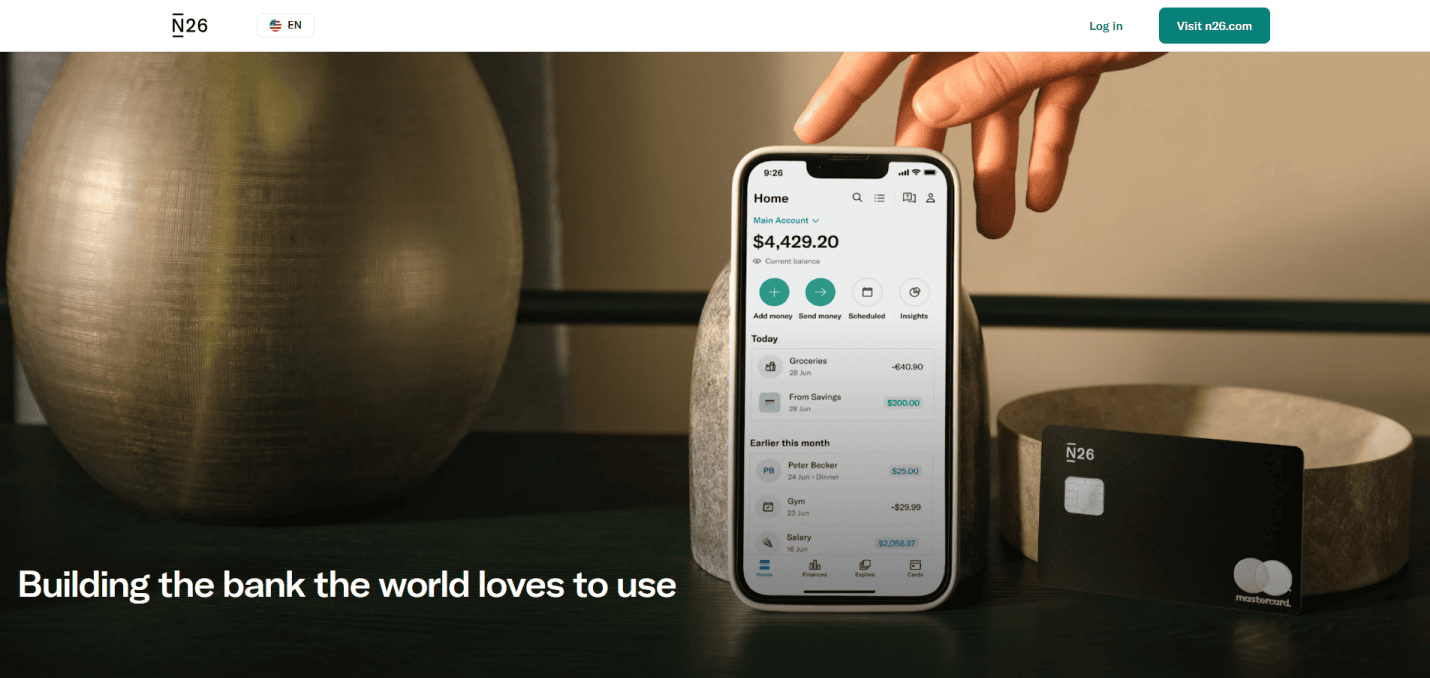 A hand holds a smartphone displaying a banking app with account details. Beside it, a black N26 Mastercard lies on a round tray. Text below reads: “Building the bank the world loves to use.”.