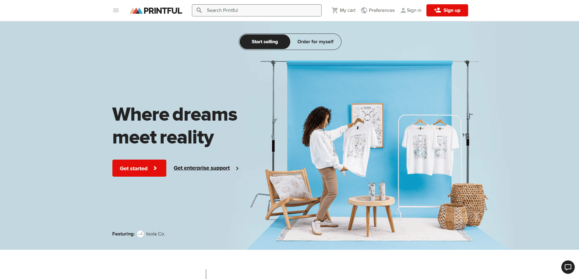 A woman holds a printed T-shirt in a bright studio with blue walls, decorated with chairs, a plant, and display racks with other printed shirts. Text reads: “Where dreams meet reality.” Buttons for getting started are visible.