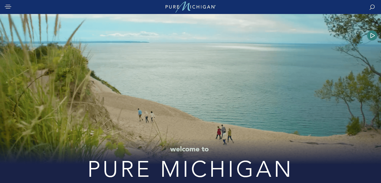 A group of people walks along a sandy beach with grassy dunes overlooking a vast lake under a partly cloudy sky. Text reads welcome to PURE MICHIGAN.