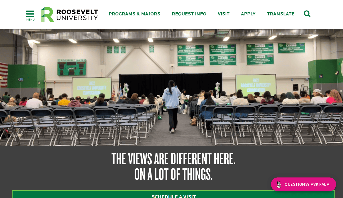 A large audience sits in rows of chairs facing a stage with two screens at Roosevelt University. Green banners hang on the back wall, and a person walks down the center aisle.