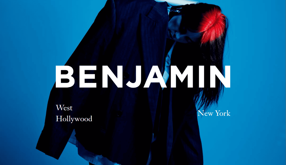 A person with black and red hair wears an oversized dark jacket against a blue background. Large white text says BENJAMIN, with West Hollywood on the left and New York on the right.