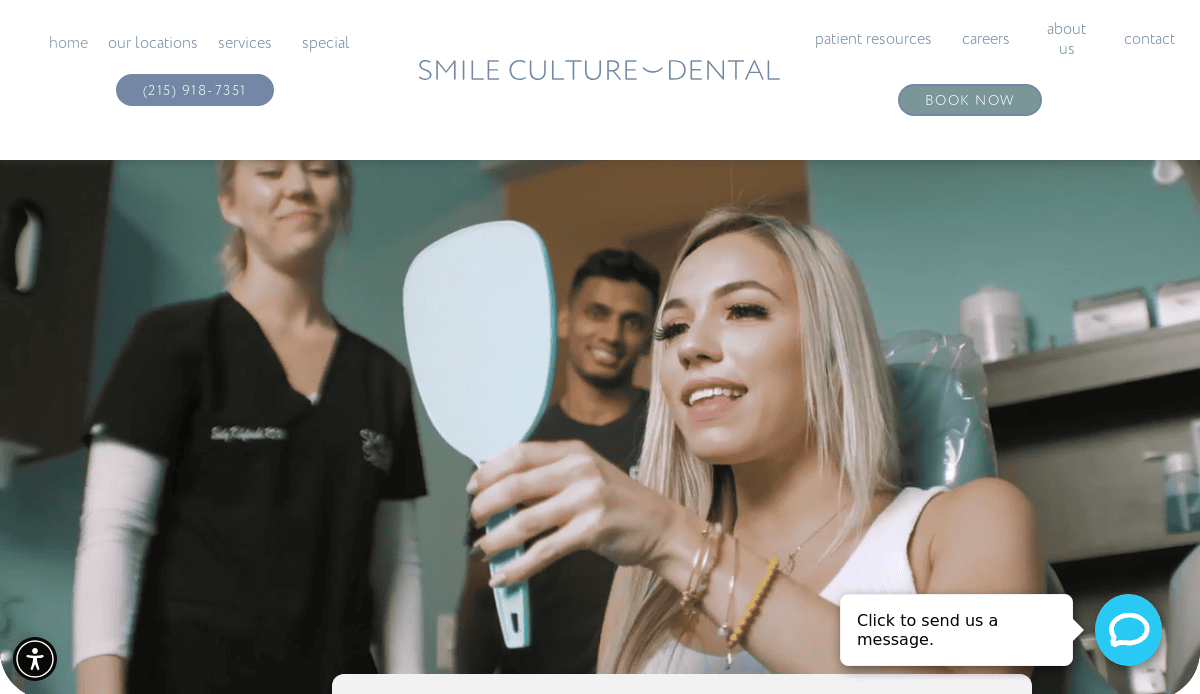 A woman sits in a dental chair smiling and holding a mirror, with two dental professionals standing nearby, all appearing happy in a modern dental office.