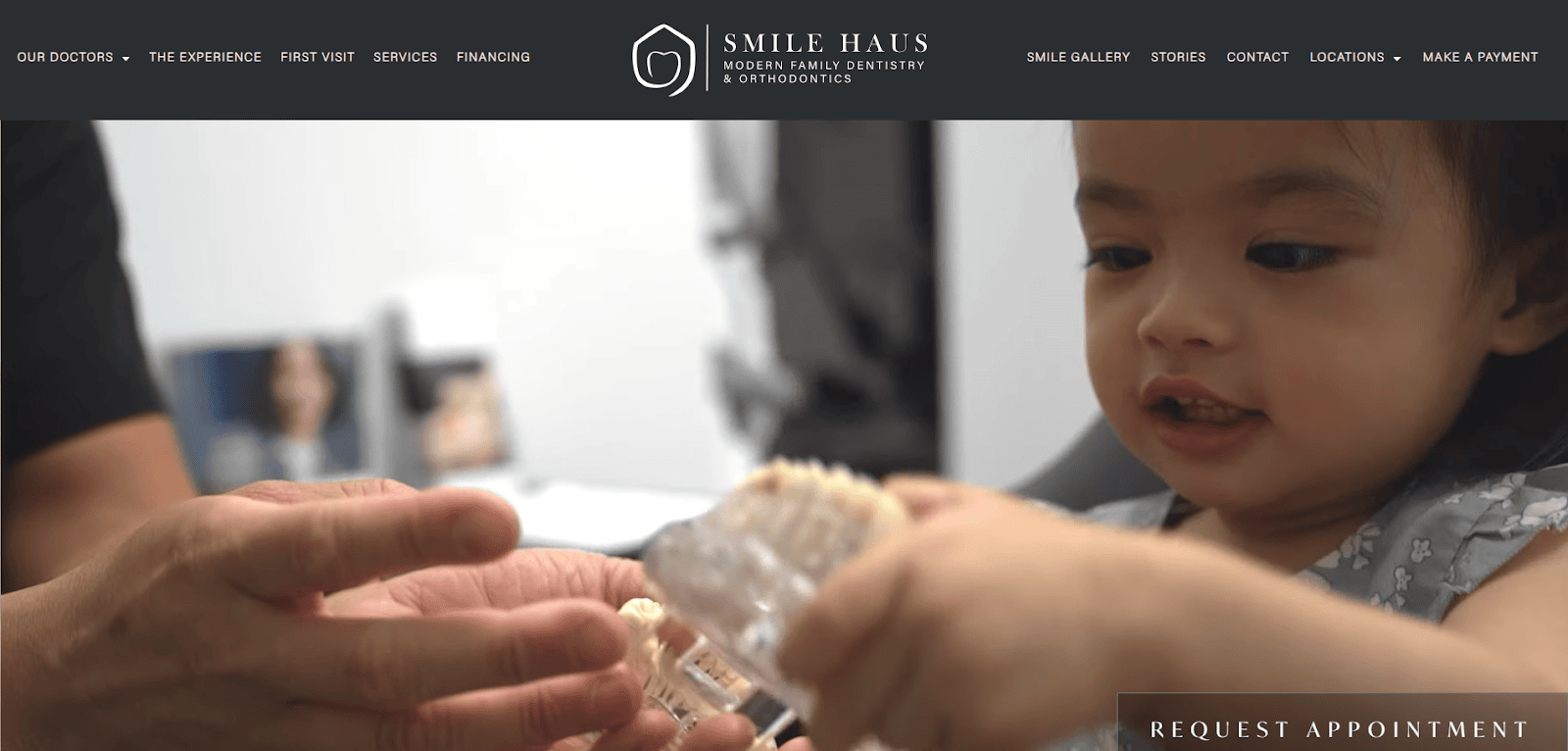 A young child examines a dental model of teeth while an adult’s hands guide her, inside a dental office. The Smile Haus dental clinic website header is visible at the top.