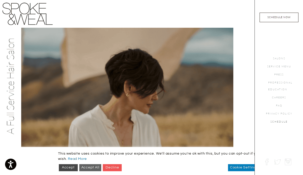 A woman with short, dark hair wearing a white top stands outdoors in a field, with hills and a beige fabric backdrop behind her. The website features navigation links and cookie consent options.