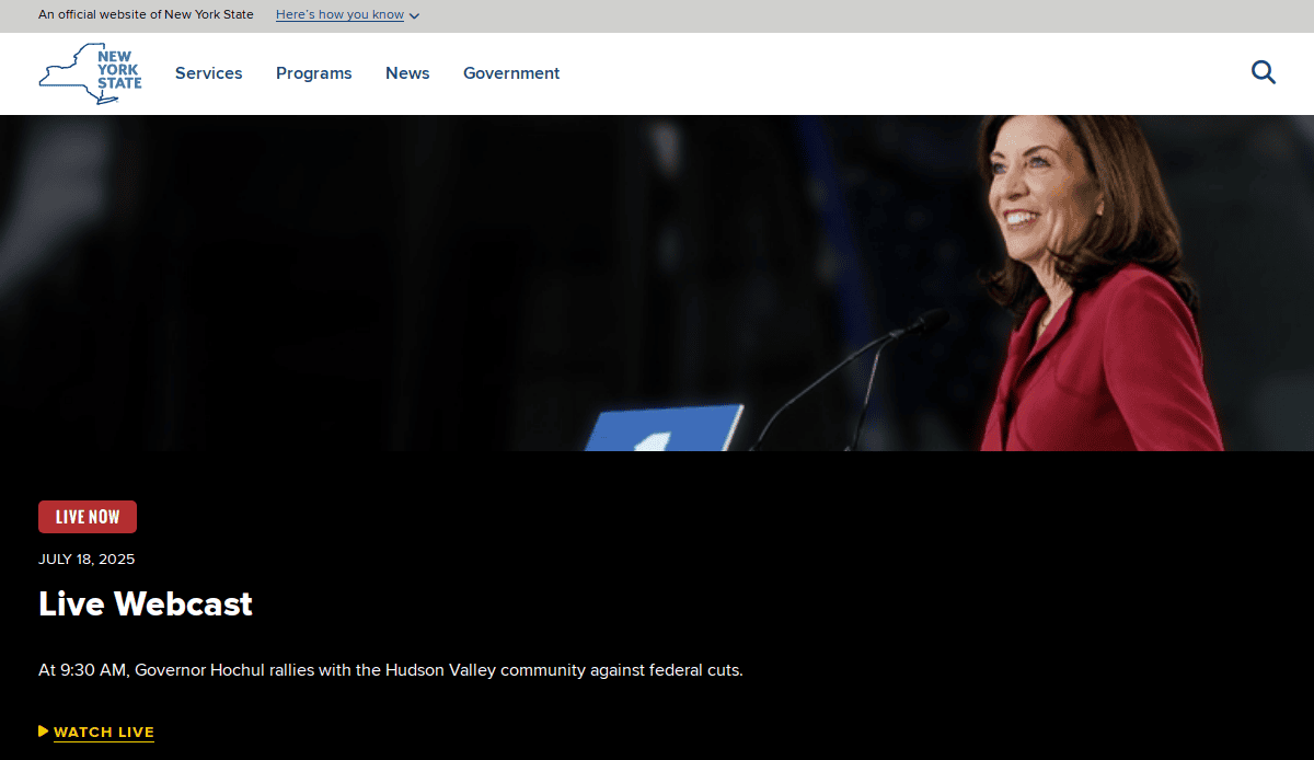 A woman in a red blazer speaks at a podium with microphones. The screen displays details for a live webcast on July 18, 2025, where Governor Hochul will rally with the Hudson Valley community against federal cuts.