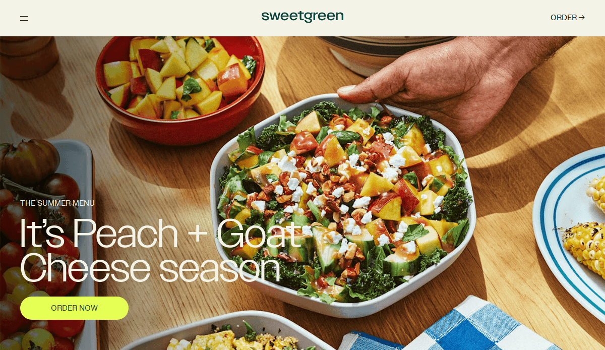 A hand holds a bowl of salad topped with peaches, goat cheese, and nuts on a wooden table with other colorful dishes. Text overlay reads: “It’s Peach + Goat Cheese season. Order now.”.