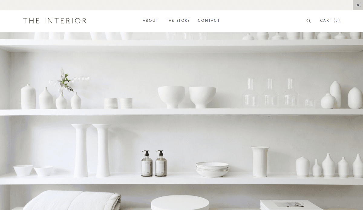Minimalist website showcasing white shelves with assorted white ceramics, glassware, and soap dispensers against a light background. Simple navigation bar with links at the top and The Interior as the site name.