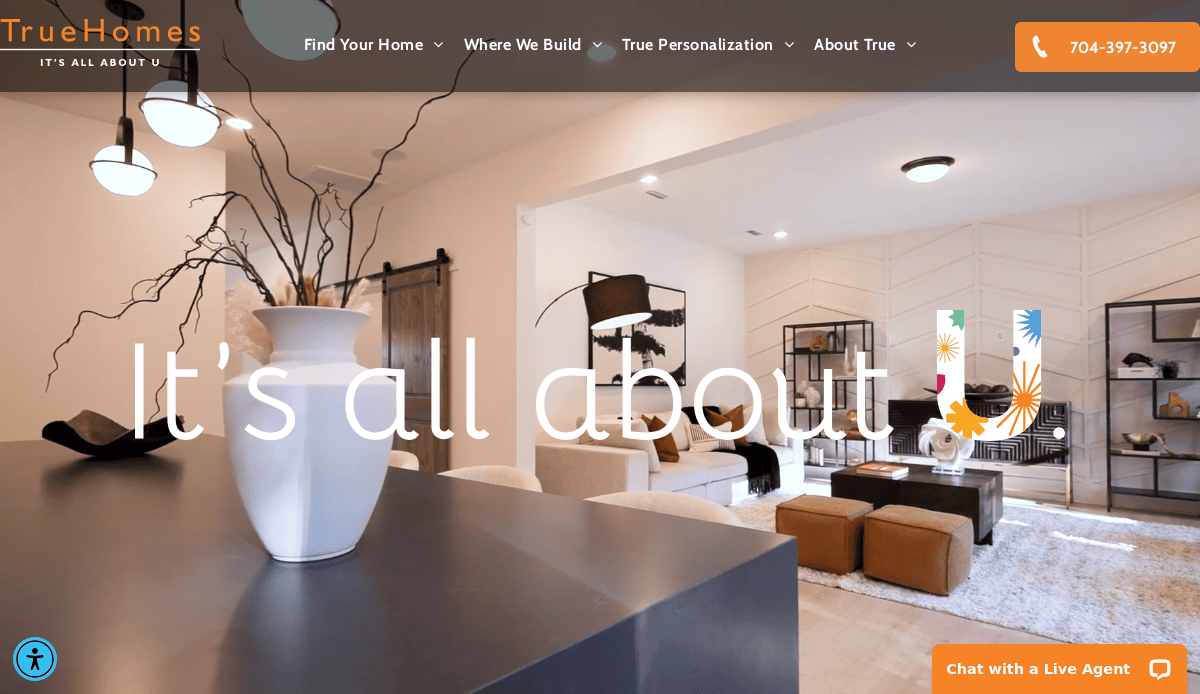 Modern living room with neutral décor, white vases on a counter, brown and white furniture, and large windows. Text overlay reads Its all about U. TrueHomes website navigation is visible at the top.