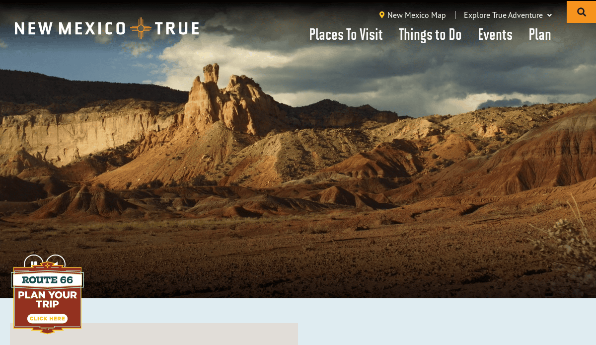 A scenic view of rugged, colorful desert cliffs and rock formations under a partly cloudy sky, featured on the New Mexico True tourism website with a Route 66 trip planning button in the lower left corner.