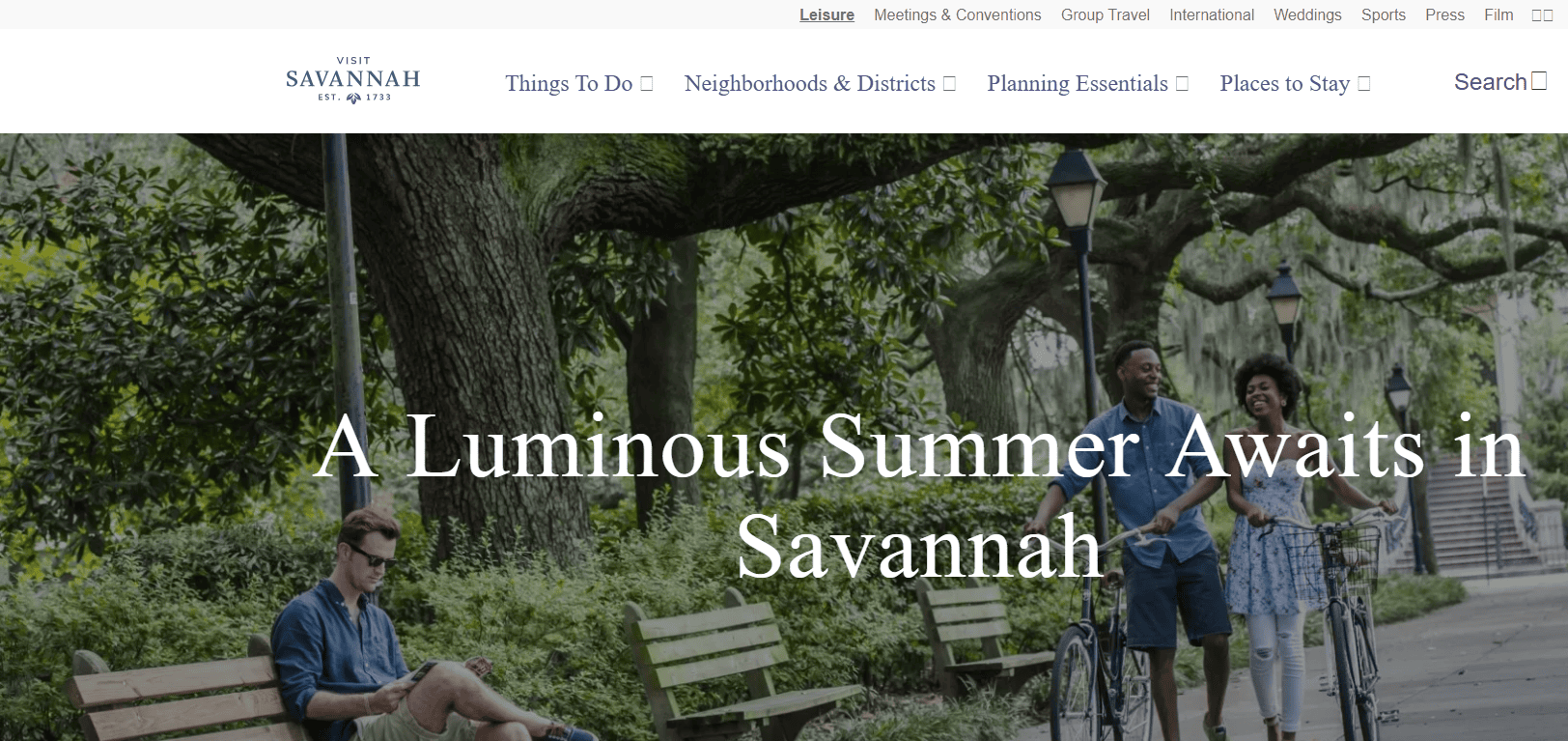 Three people enjoy a park in Savannah; one sits on a bench, and two others walk and ride bikes under large trees draped with Spanish moss. Text reads: “A Luminous Summer Awaits in Savannah.”.