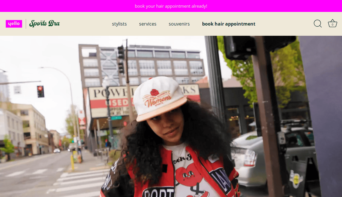 A person with long curly hair wearing a graphic jacket and a white cap walks on a city street. Buildings, trees, cars, and a traffic light are visible in the background.